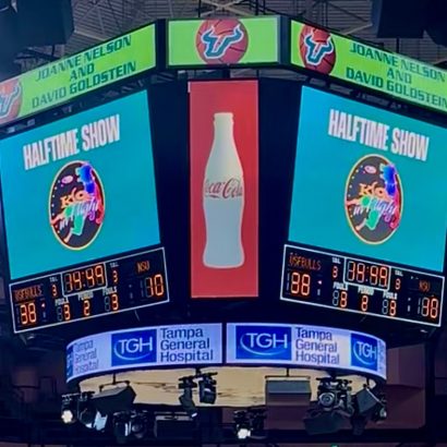 Scoreboard displays 'HALFTIME SHOW' with a Coca-Cola bottle ad in the center and event banners overhead, in a sports arena.