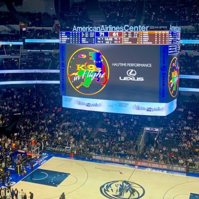 Basketball arena full of fans with a towering video scoreboard showing a halftime performance graphic and Lexus logo above the court.