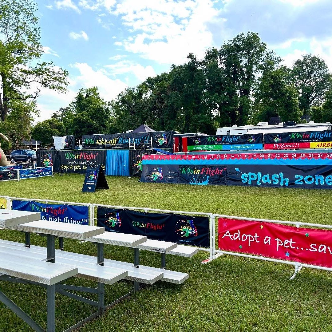Outdoor dog-themed event setup with colorful banners reading K9s in Flight and a large inflatable dog mascot on the left; bleachers in the foreground.