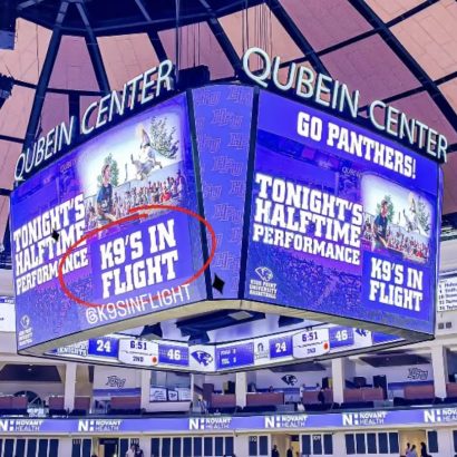 Jumbo scoreboard in a sports arena showing a promo graphic 'K9's in Flight' circled, with 'Tonight's Halftime Performance' text nearby, at Qubein Center.