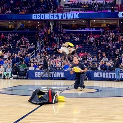 Georgetown arena performance: a man kneels on the court tossing yellow frisbees as a bag of discs lies on the floor, with spectators in the stands.