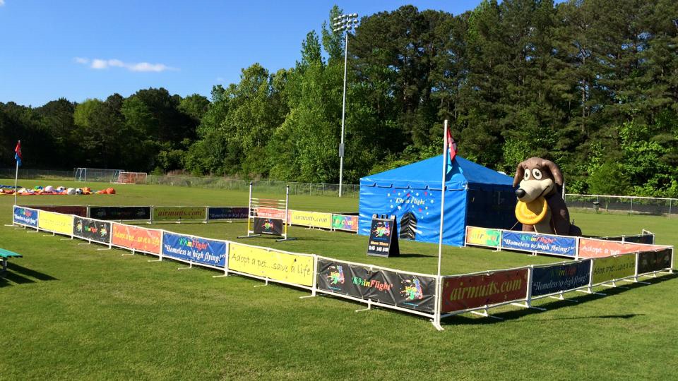 Outdoors festival area with colorful banners around a fenced play zone, a blue tent, and a large inflatable cartoon dog ponytail or ring beside it.